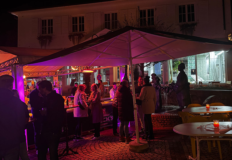 Mehrere Grüppchen Partygäste stehen im Dunkeln an einem beleuchteten Bierwagen und unterhalten sich.
